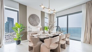 Un espace repas moderne avec une grande table dressée pour six personnes, entourée de six chaises. Un pot de fleurs se trouve à gauche, et une grande fenêtre offre une vue sur le paysage urbain et la mer.