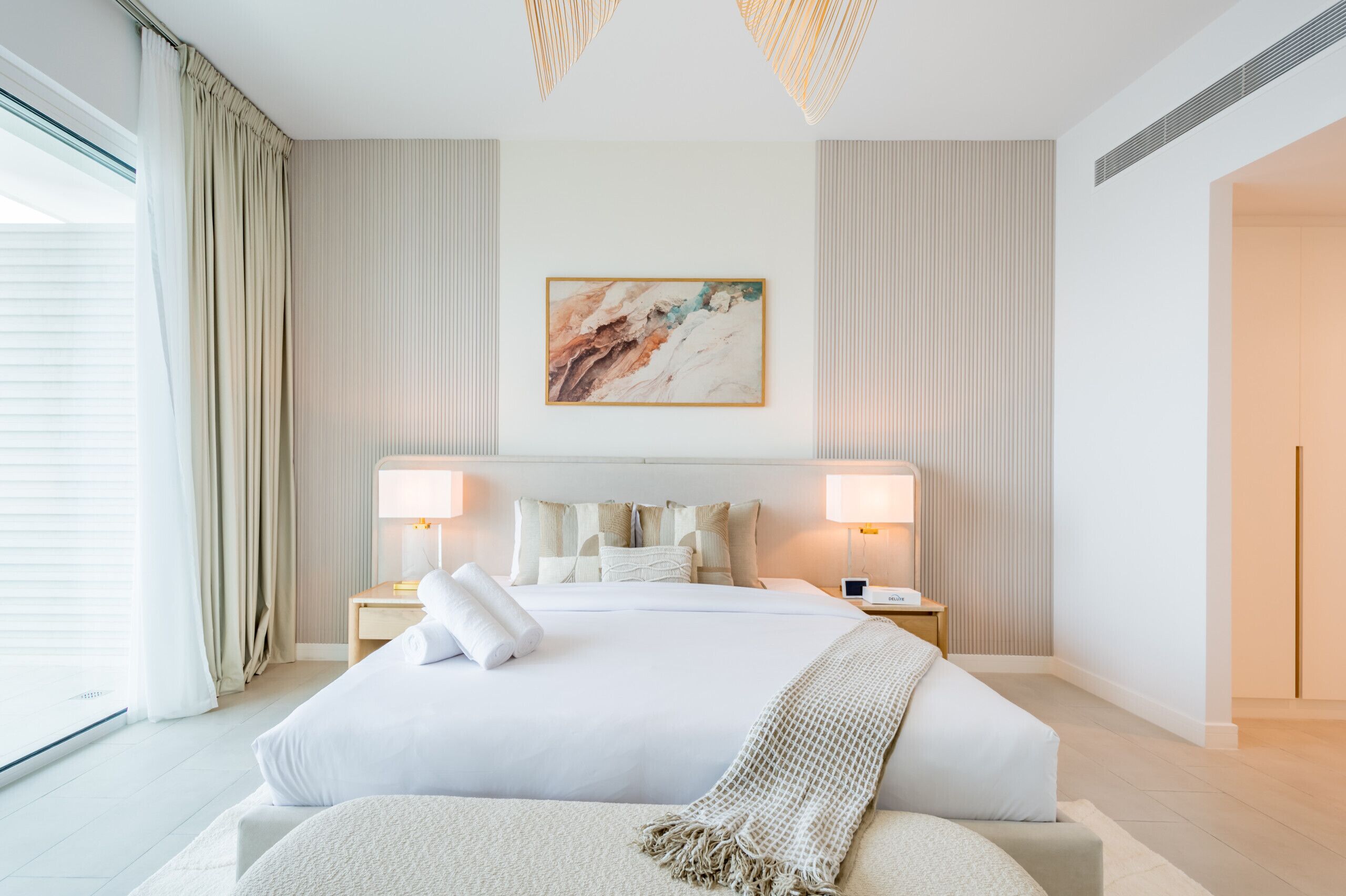 A modern hotel room with a large bed, two bedside lamps, a painting on the wall, and a view of the outdoors.