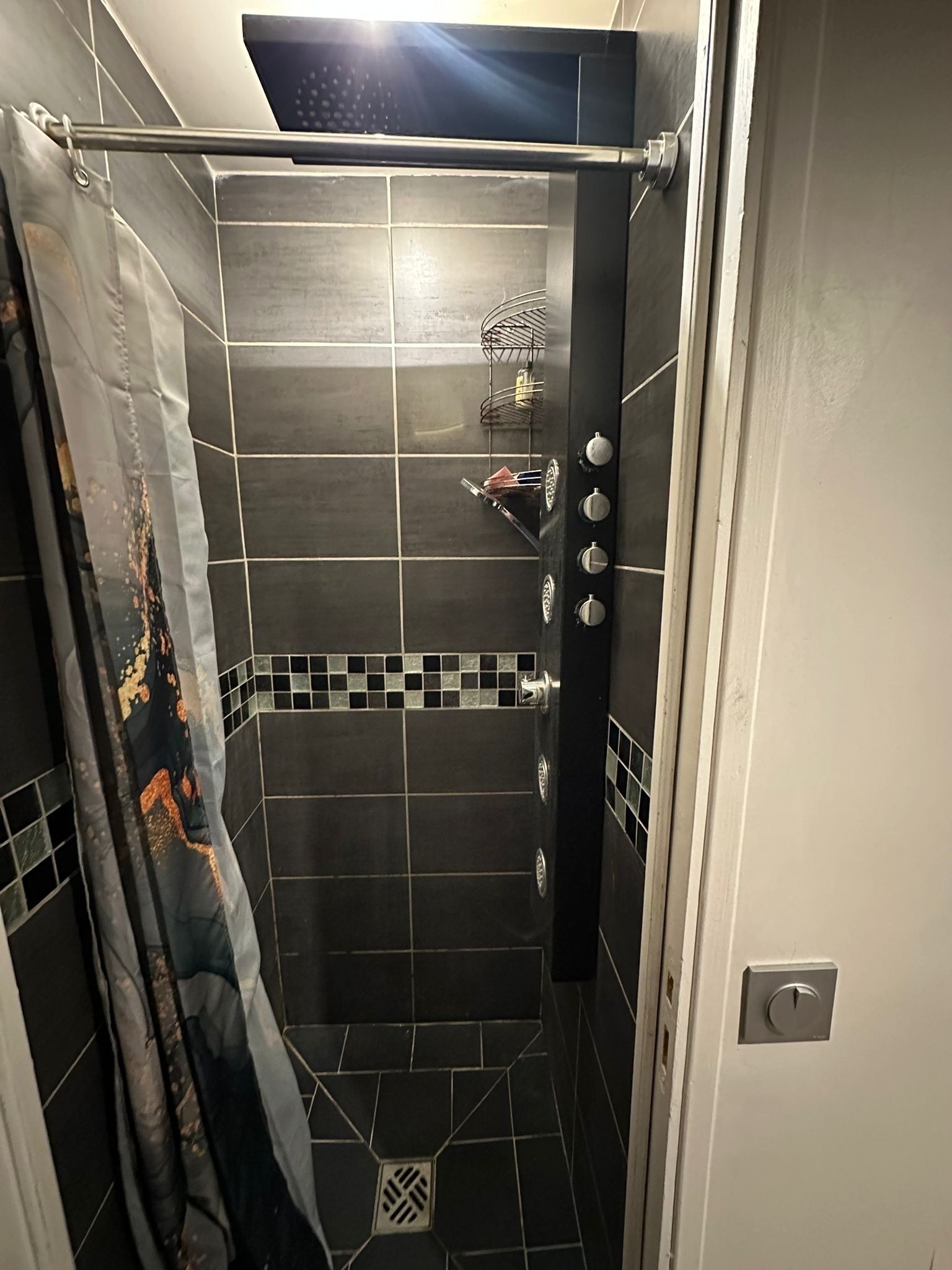Une douche avec un rideau de douche en métal, une étagère avec des articles de toilette et un sol carrelé.