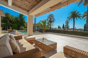 A covered outdoor seating area with wicker furniture, a glass table, and a pool in the background.