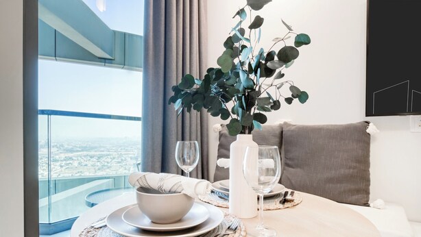 A dining area with a table set for two, a vase with green foliage, and a city view through large windows. - Luxury Skyline Studio | Pool and Stunning Views (Dubai)