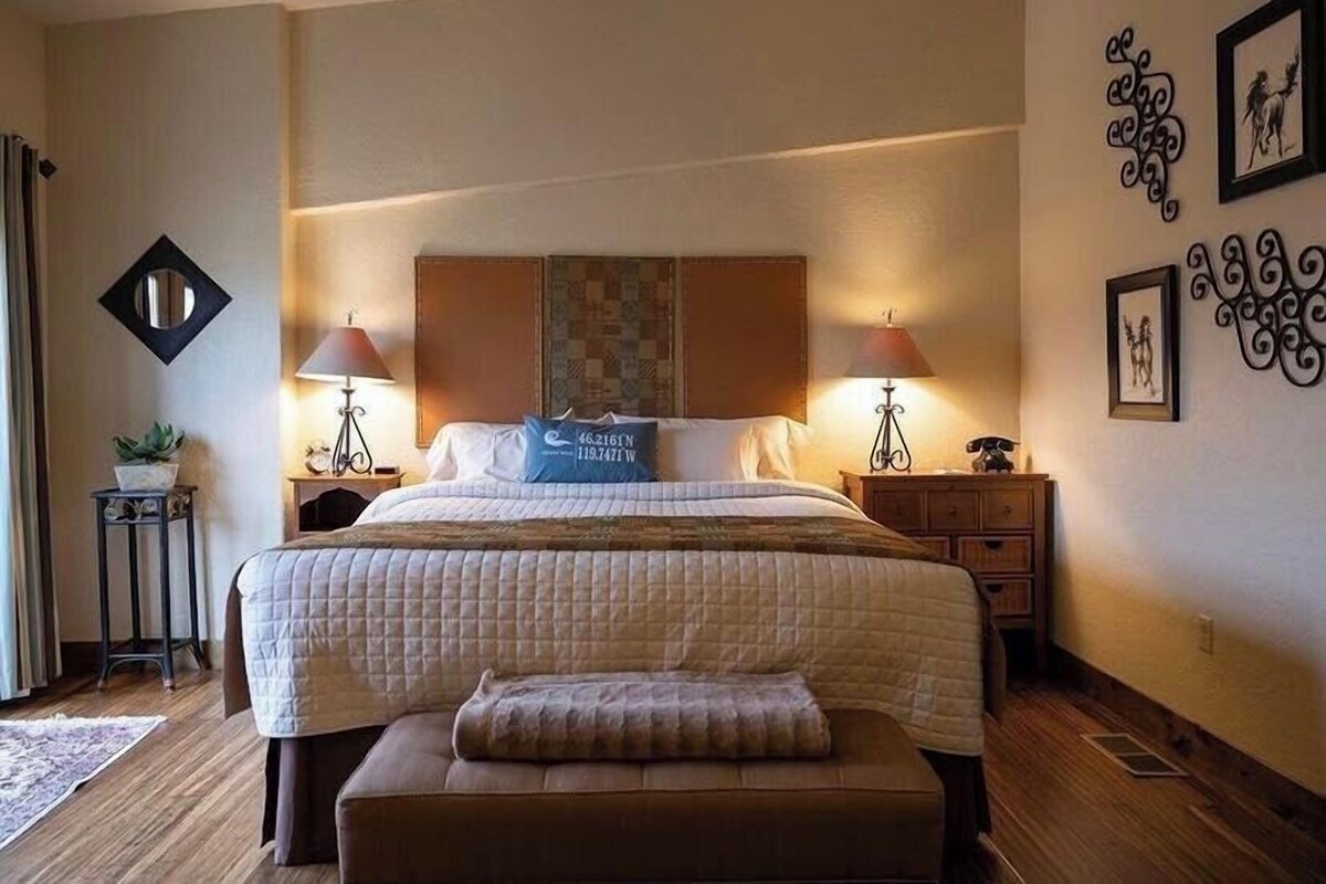 A hotel room with a large bed, two bedside tables, a nightstand, a decorative wall piece, a small potted plant, and a window with curtains.