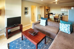 A living room with a sofa, coffee table, and TV. There is a kitchen in the background with wooden cabinets and a dining area.