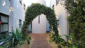 Un estrecho sendero de ladrillo bordeado de exuberante vegetación y un arco cubierto de hiedra que conduce a un edificio.