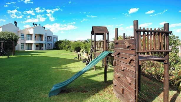 Un parque infantil con un tobogán y estructuras para escalar, rodeado de un césped cuidado y un edificio al fondo.