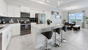 A modern kitchen with white cabinets, stainless steel appliances, a central island with bar stools, and a dining area with a view.