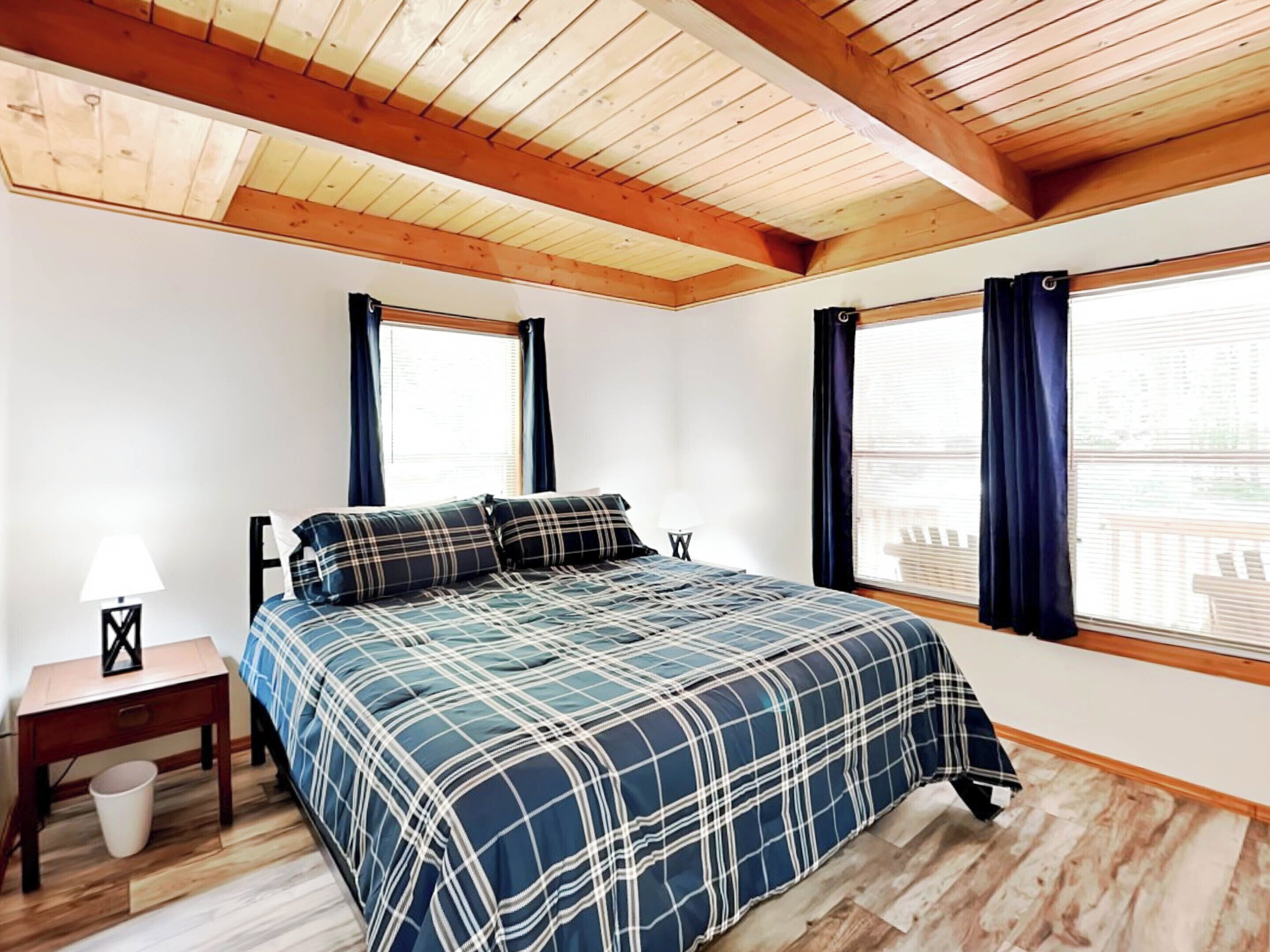 Une chambre avec un plafond en bois, deux fenêtres avec des rideaux, un lit recouvert d’une couverture à carreaux, une table de chevet et une lampe.