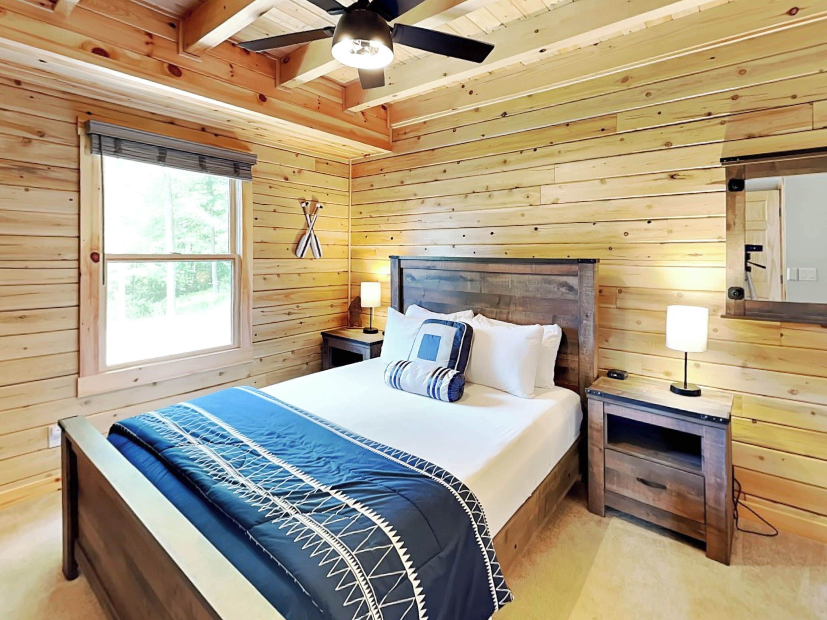 A wooden cabin bedroom with a bed, nightstands, a ceiling fan, and a window.