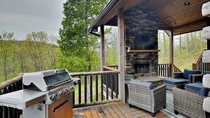 A covered outdoor deck with a barbecue grill, wicker furniture, and a stone fireplace. - Sunset Bluff by Taylor Made Deep Creek (McHenry)