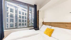 Ein Hotelzimmer mit großem Fenster, einem Bett mit weißer Bettwäsche und einem gelben Kissen sowie einem Holz-Kopfteil.