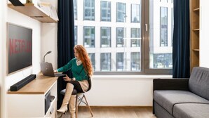Eine Person sitzt an einem Schreibtisch mit einem Laptop, einem Fernseher, der 'NETFLIX' zeigt, und einem Fenster mit Vorhängen.