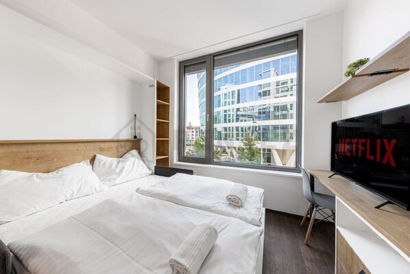 A modern hotel room with a large bed, a desk with a TV, and a window with a city view.