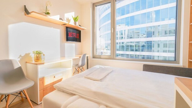 Ein modernes Hotelzimmer mit einem großen Bett, einem Schreibtisch mit Stuhl, einem an der Wand befestigten Fernseher und einem großen Fenster mit Blick auf die Stadt.