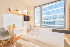 A modern hotel room with a large bed, a desk with a chair, a TV mounted on the wall, and a large window offering a city view.