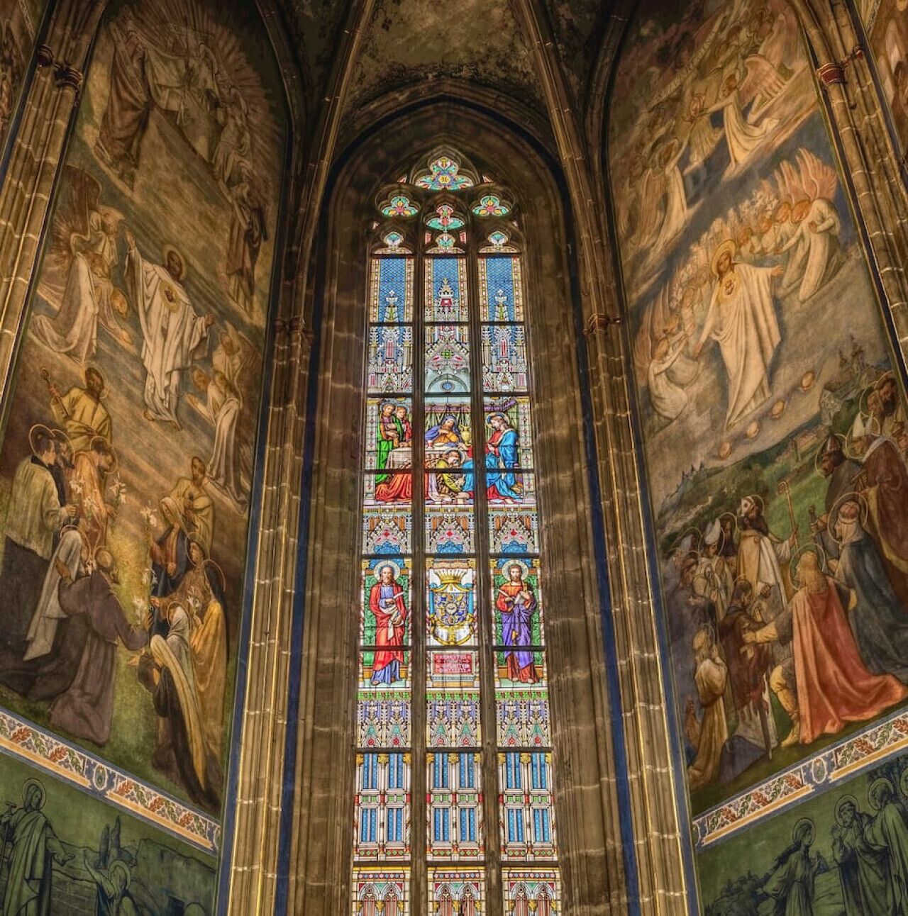 A stained glass window with intricate designs and figures, set within a vaulted ceiling with detailed frescoes.