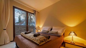 A neatly made bed with a patterned blanket and pillows, a bedside lamp, and a view of a balcony with a railing.