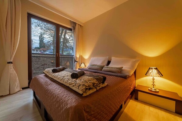 A neatly made bed with a patterned blanket and pillows, a bedside lamp, and a view of a balcony with a railing.