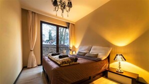 A bedroom with a large bed, a bedside table with a lamp, and a view of the outside through a window.