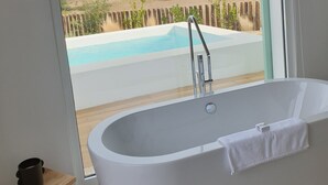 Ein modernes Badezimmer mit einer freistehenden Badewanne, einem hölzernen Hocker und Blick auf einen Pool und die Landschaft.