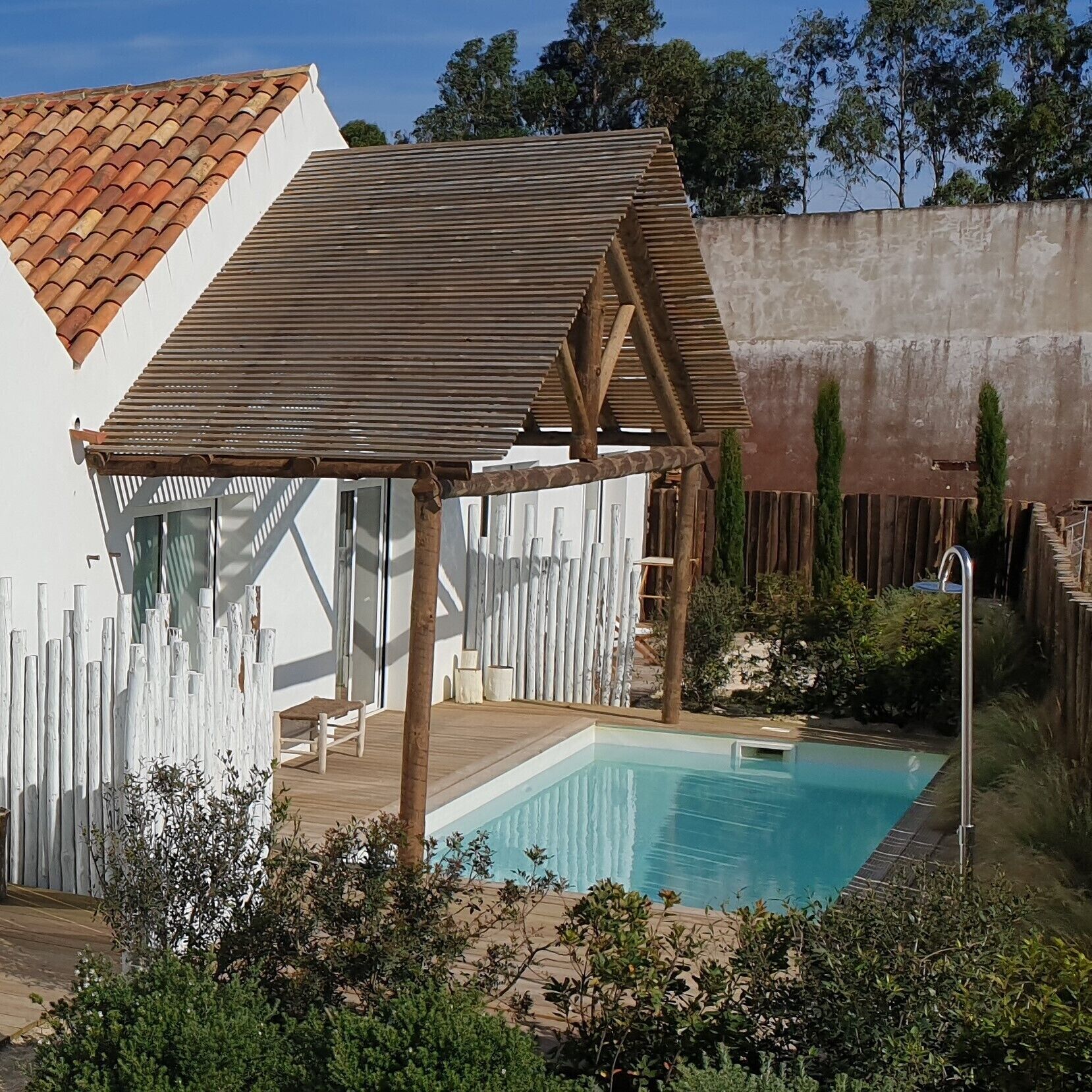 A pool area with a wooden pergola, a white fence, and a view of a house with a tiled roof.