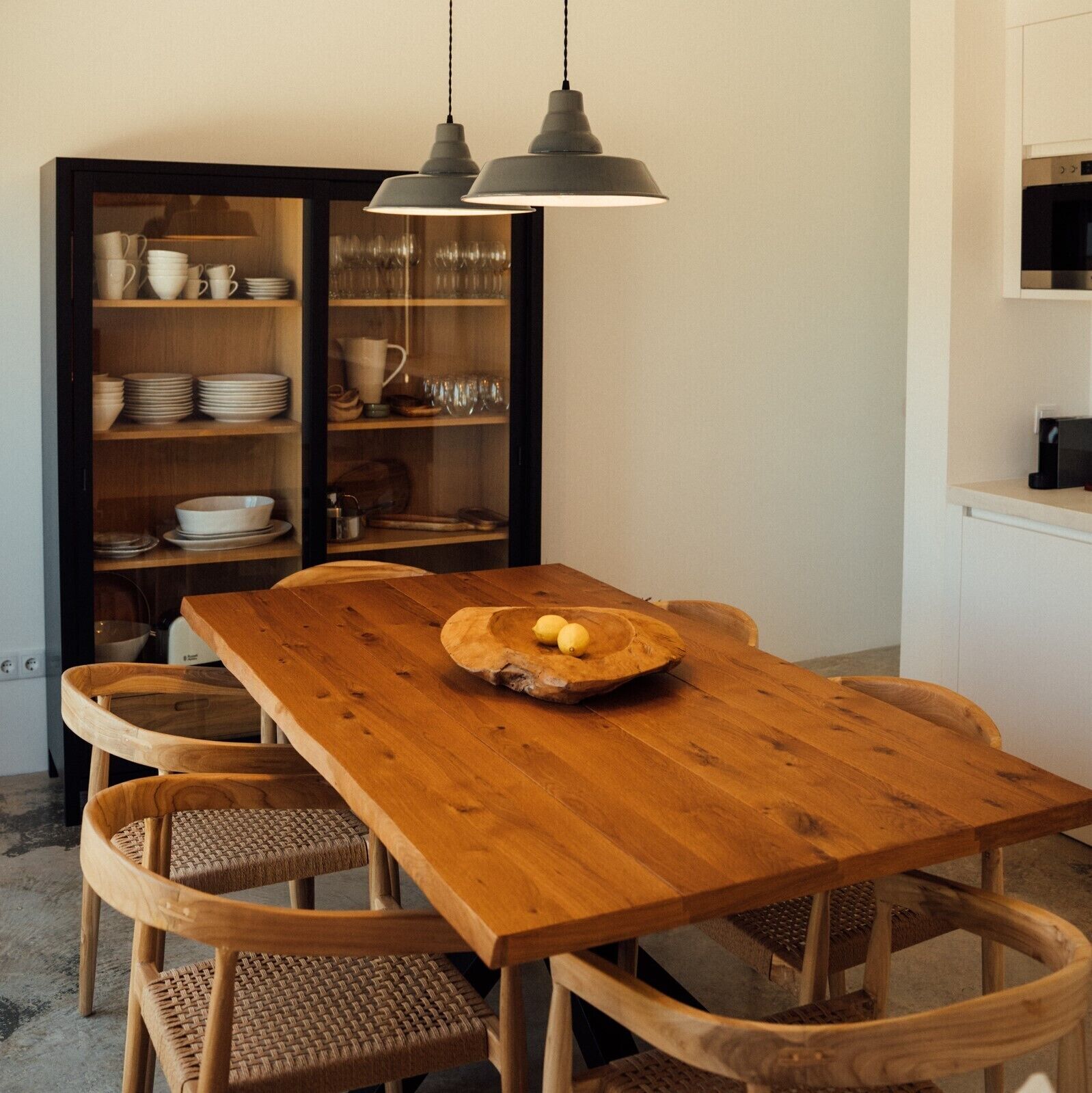 A wooden dining table with a woven chair, a glass cabinet with dishes, and pendant lighting.
