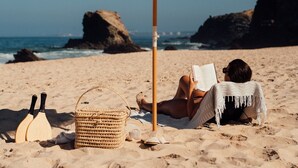 Eine Person entspannt sich unter einem Strandsonnenschirm an einem sandigen Strand, im Hintergrund liegen Felsen und das Meer.