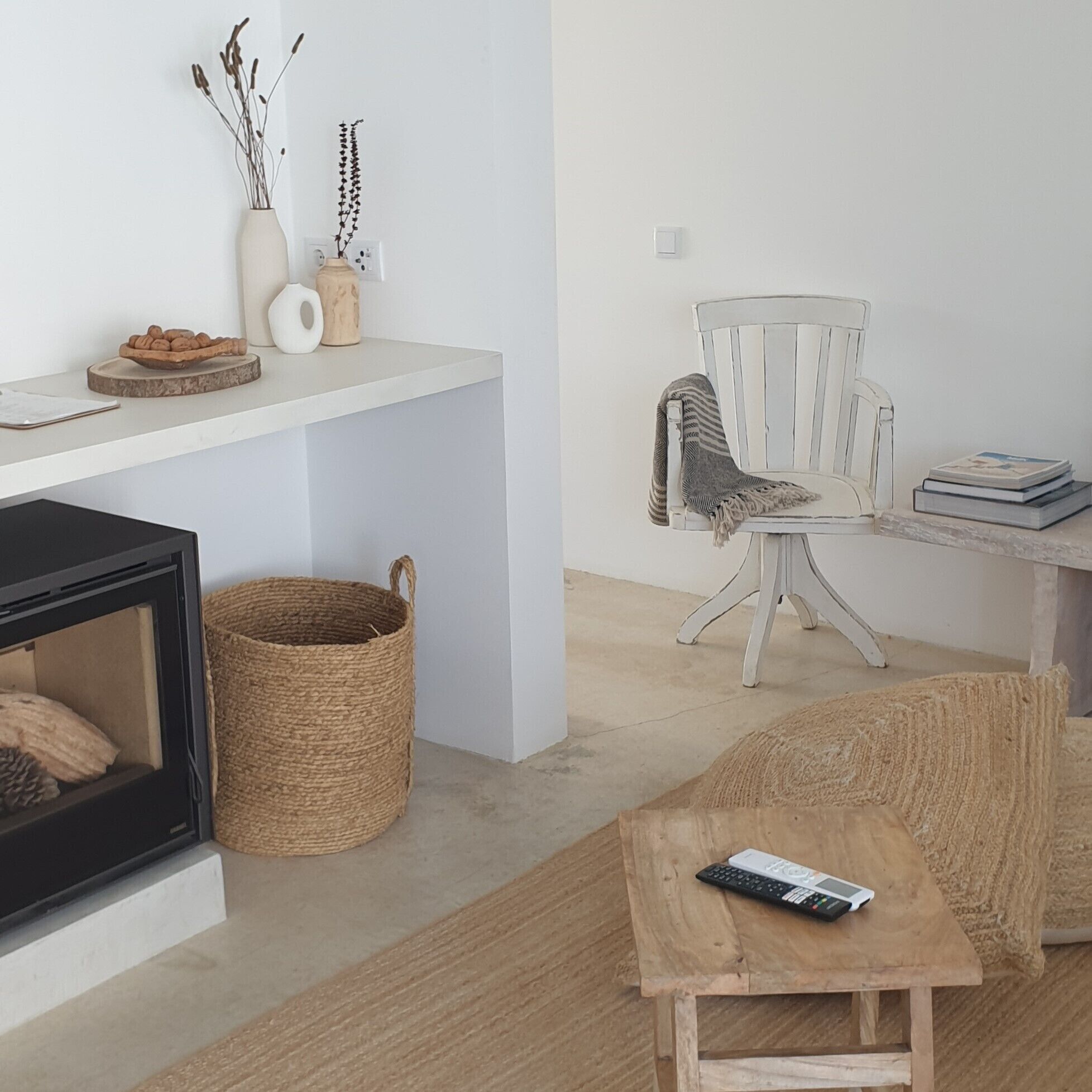 A living room with a fireplace, a woven basket, a small wooden table with a remote control, a striped chair, and a white shelf with decorative items.