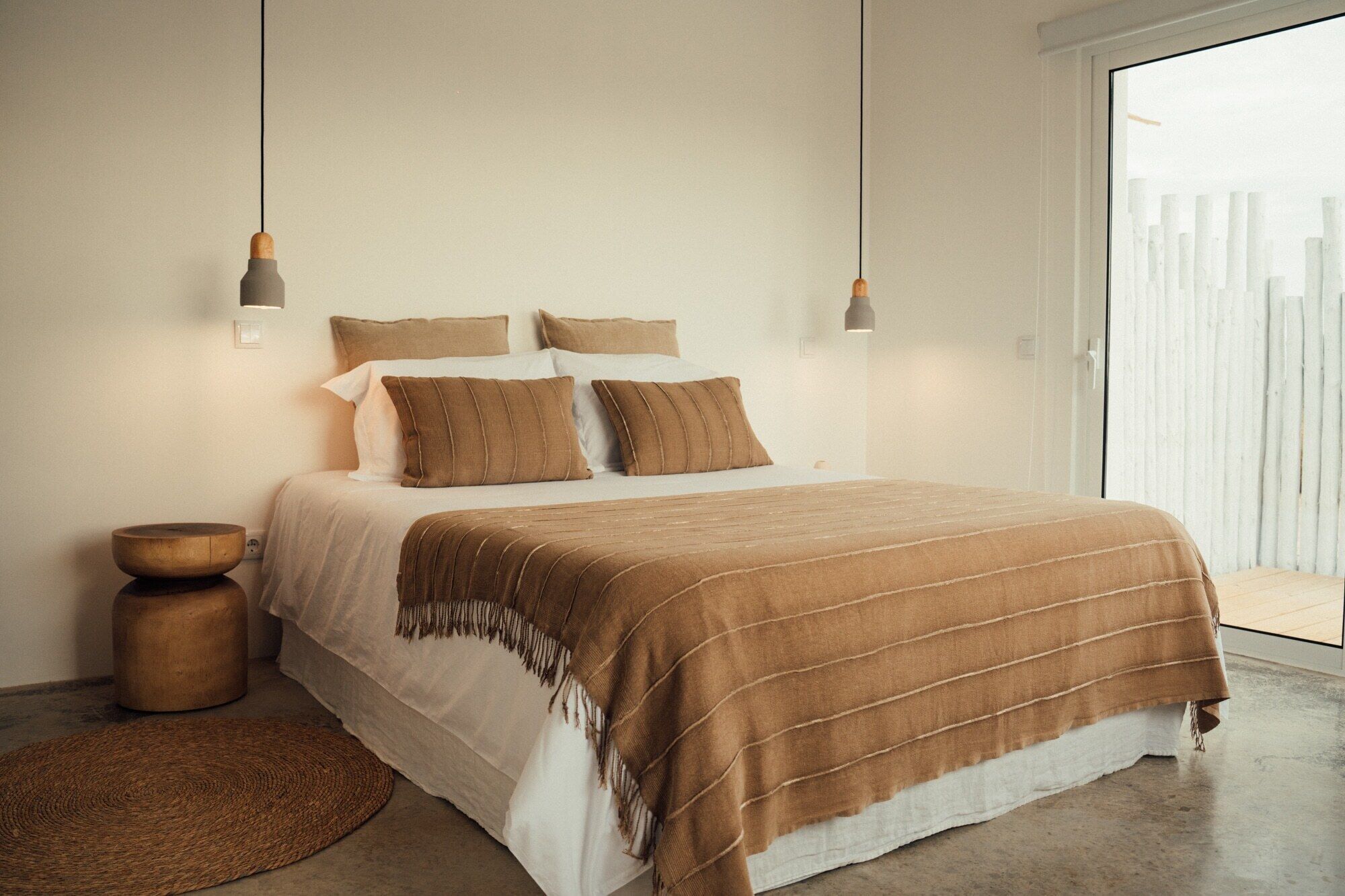 A modern bedroom with a large bed, a wooden side table, a woven rug, and a view of a white fence through a large window.