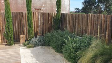 A sandy garden area with a wooden fence, surrounded by various shrubs and plants.