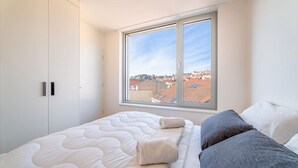 Une chambre avec une grande fenêtre, un lit doté d’un matelas blanc et d’une couverture grise, et des murs blancs.