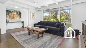A modern living room with a dark grey sofa, a wooden coffee table, and a patterned rug. There are large windows offering a view of greenery outside. - Blairgowrie Back Beach Holiday Escape (Blairgowrie)