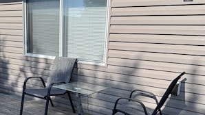 A covered outdoor seating area with two chairs and a table, adjacent to a house with a window and a wall-mounted light fixture. - Cozy and Charming Amber Valley Haven in Fargo (Fargo)