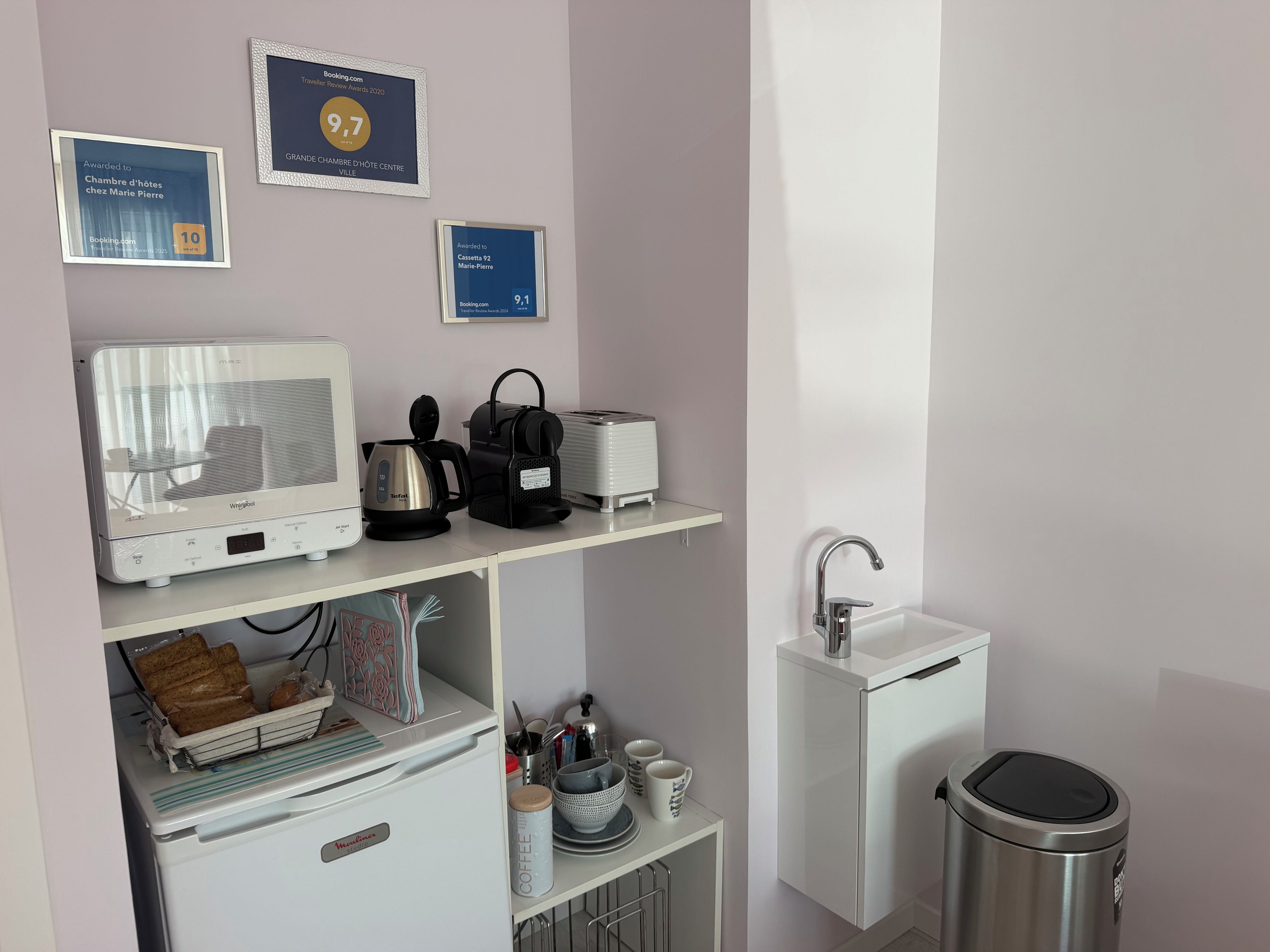 Una kitchenette con microondas, tostadora, hervidor de agua y cafetera sobre una encimera blanca. En la pared, encima de la encimera, hay cuadros con reconocimientos.