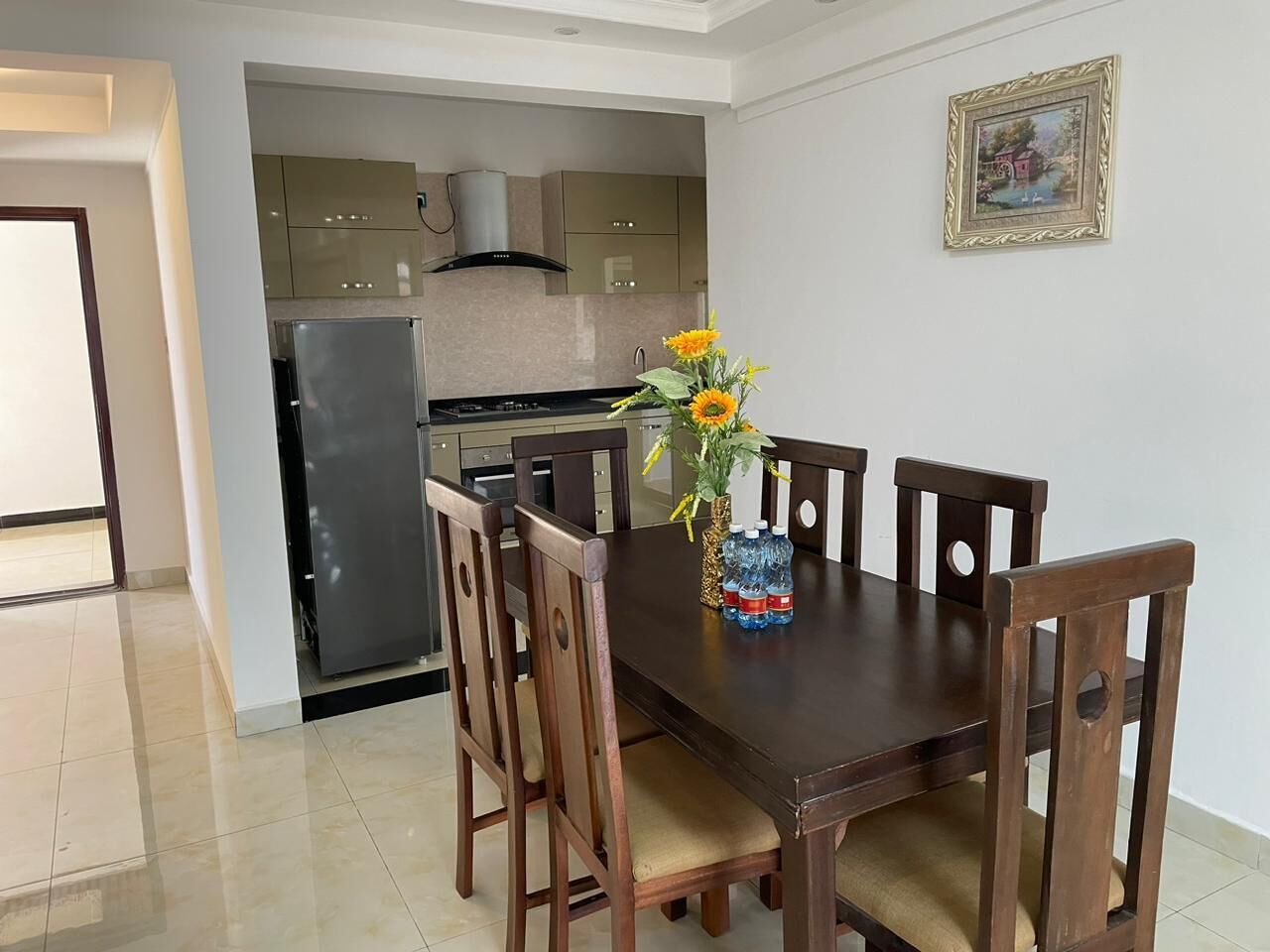 A dining area with a dark wooden table and chairs, a vase with flowers, and a kitchen in the background.