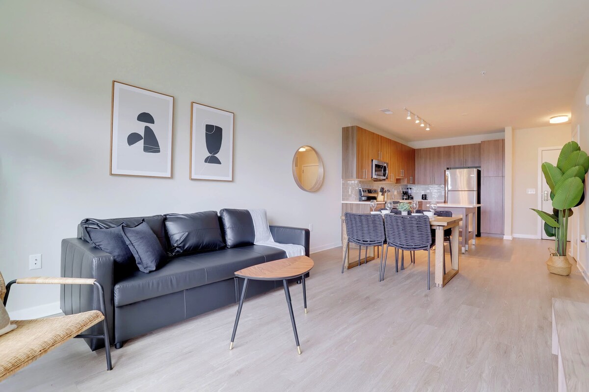 A modern living room with a grey sofa, a small wooden table, and a dining area with chairs and a kitchen in the background.