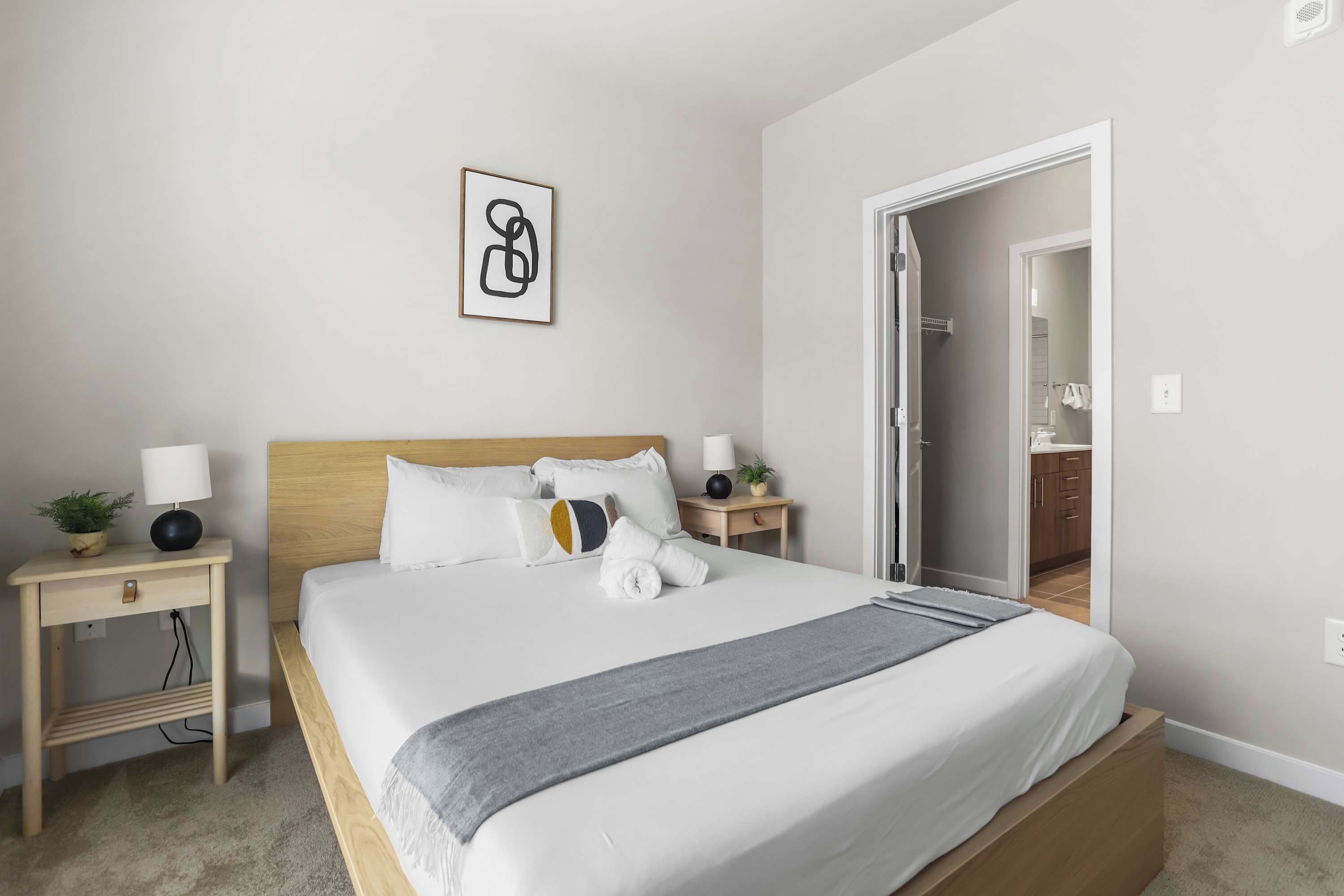 A neatly arranged bedroom with a bed, bedside tables, a nightstand, a wall-mounted artwork, and an open door leading to a bathroom.