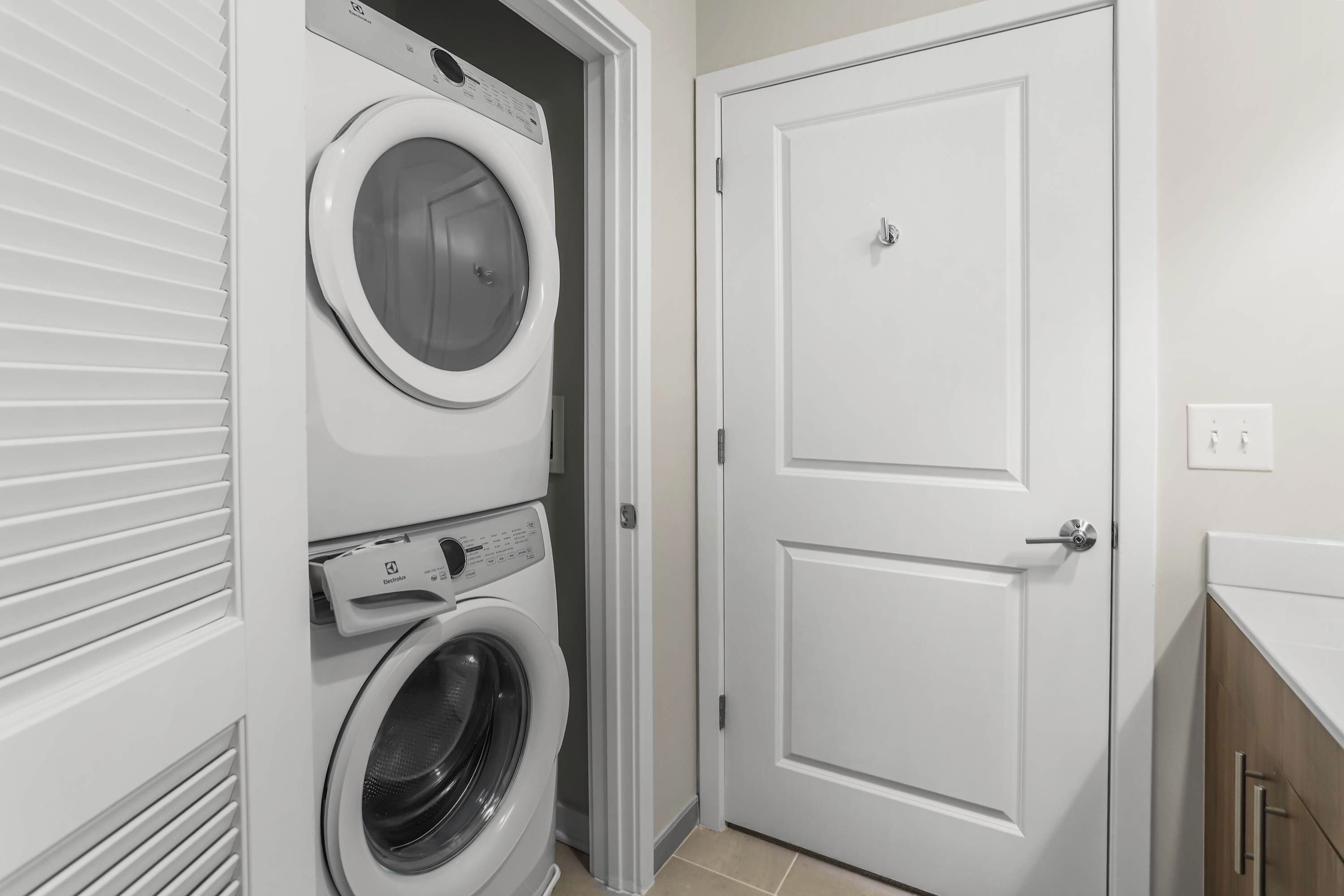 A laundry room with a front-loading washer and dryer, a door with a peephole, and a light switch on the wall.