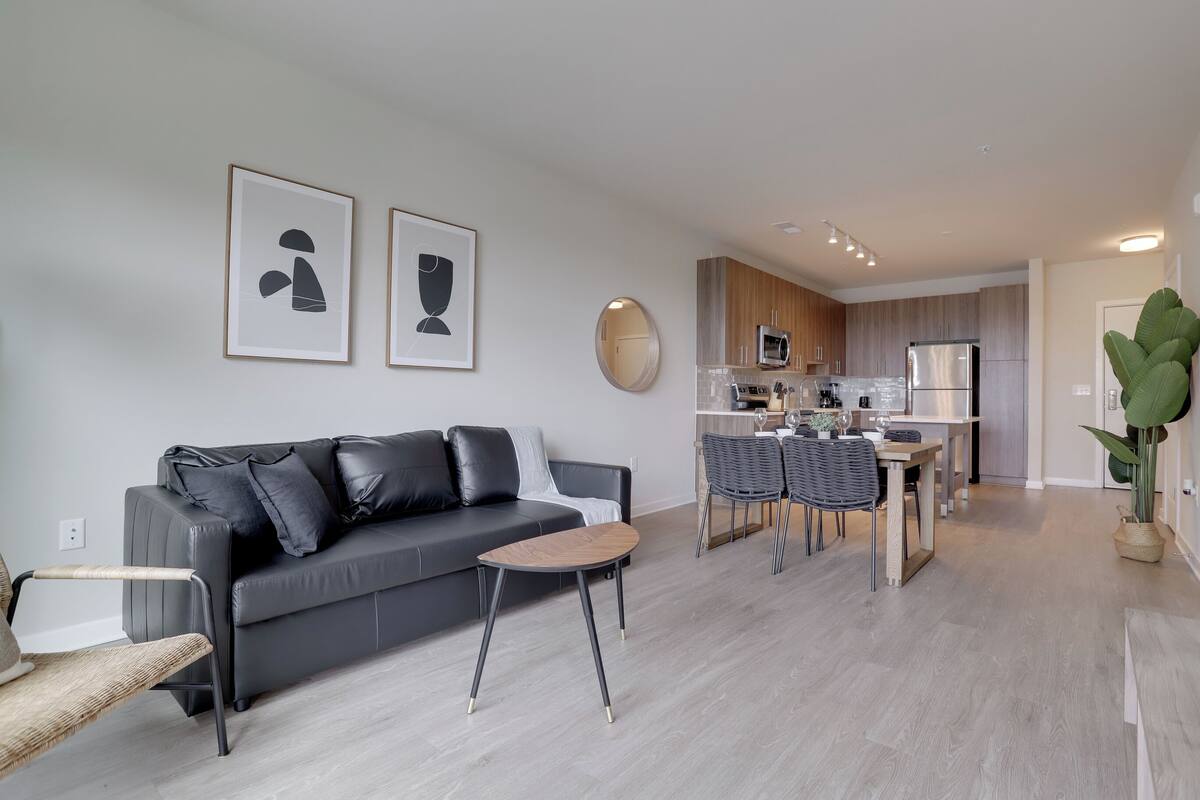 A modern living room with a grey sofa, a small wooden coffee table, and a dining area with a kitchen in the background.