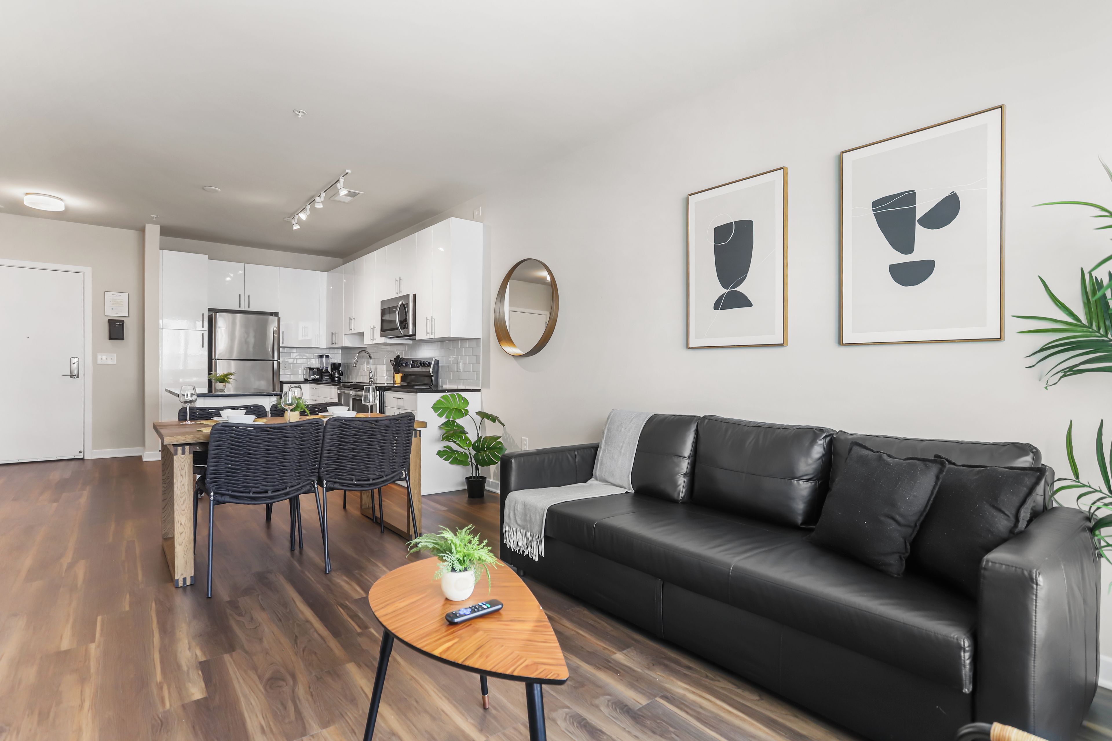 A modern living room with a black leather sofa, a wooden coffee table, and a dining area with wicker chairs. There are two framed abstract paintings on the wall.