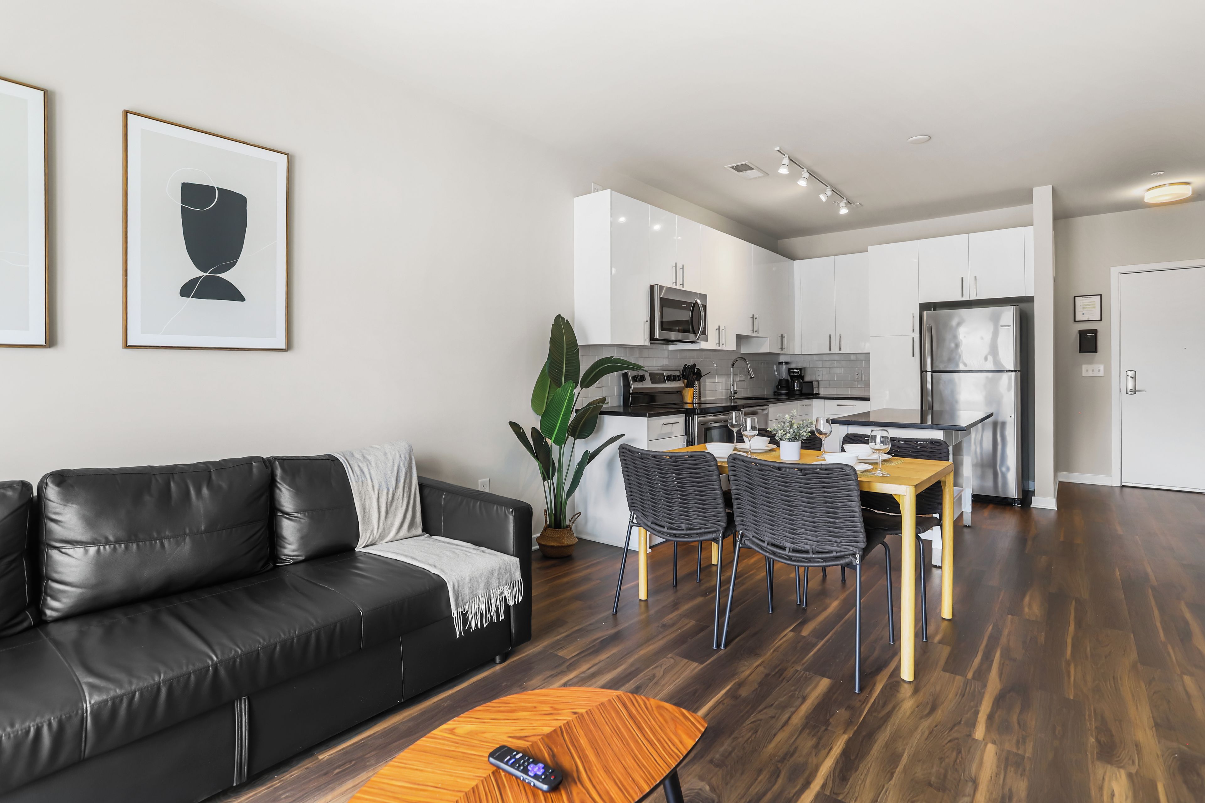 Una sala moderna con un sofá de cuero negro, una mesa de comedor con sillas y una zona de cocina con armarios blancos y electrodomésticos de acero inoxidable.