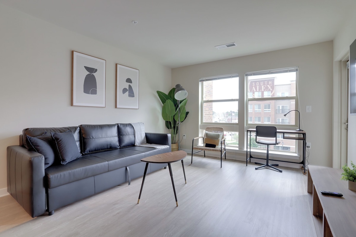 Una sala moderna con un sofá gris, una mesita de madera y una silla de oficina negra. Se ven dos ventanas con vistas a edificios.