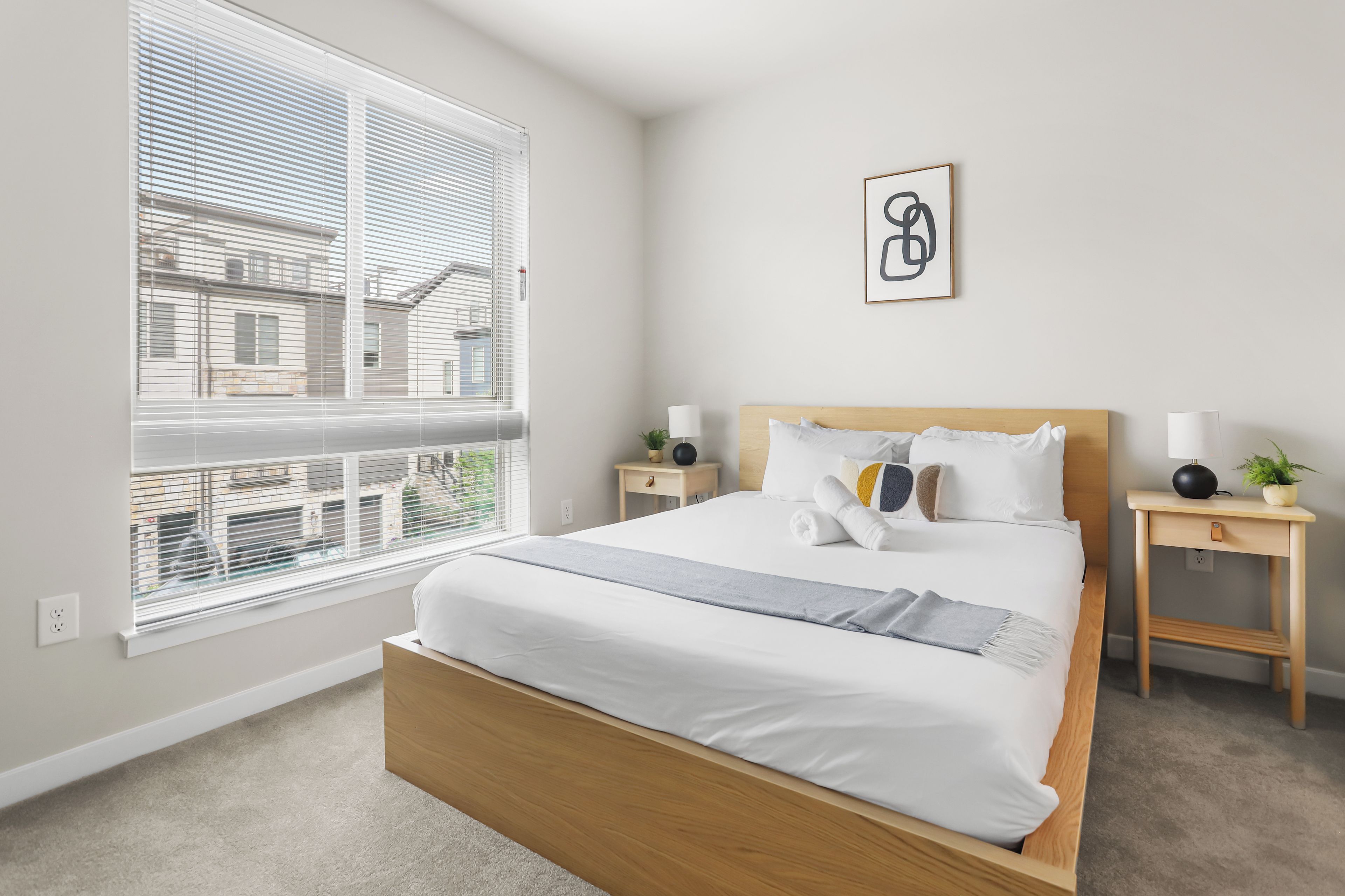 A hotel room with a bed, bedside tables, a lamp, a plant, and a window with blinds.