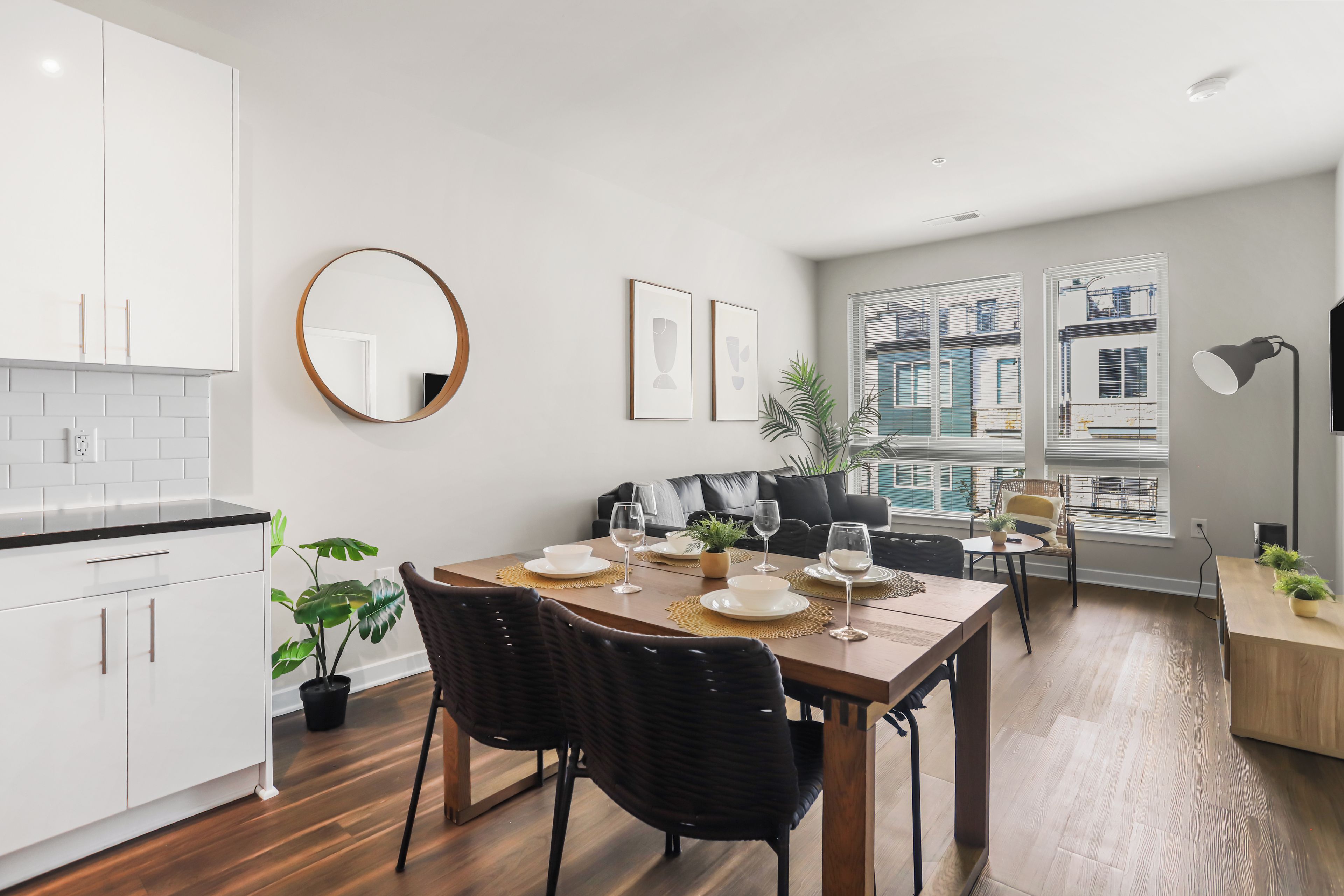 A modern kitchen with white cabinets, a dining area with a wooden table set for four, and a large window with blinds.
