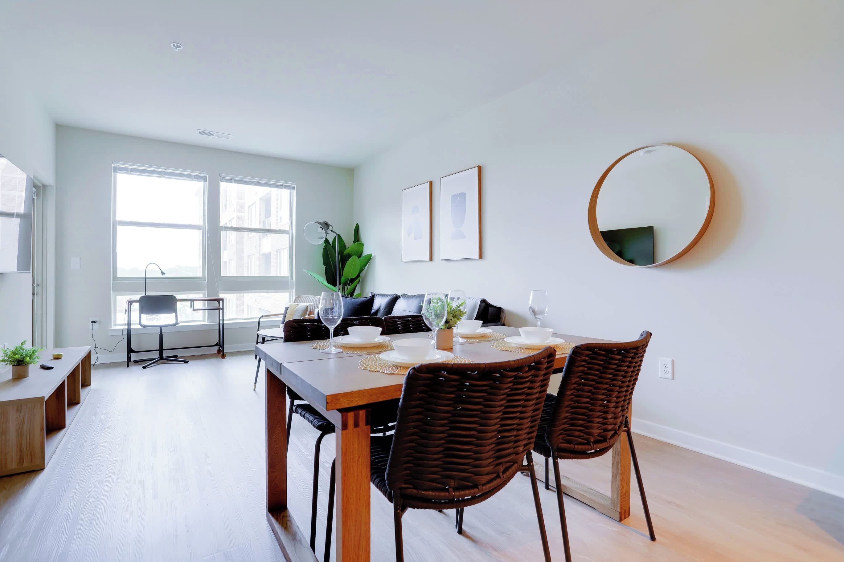 Un comedor con una mesa de madera dispuesta para cuatro personas, rodeada de sillas de mimbre, un sofá de cuero negro y una planta en maceta.