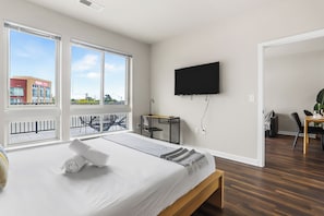 Moderni hotellihuone, jossa on suuri sänky, työpöytä, televisio ja parveke, jolta on näkymä kaupunkiin.