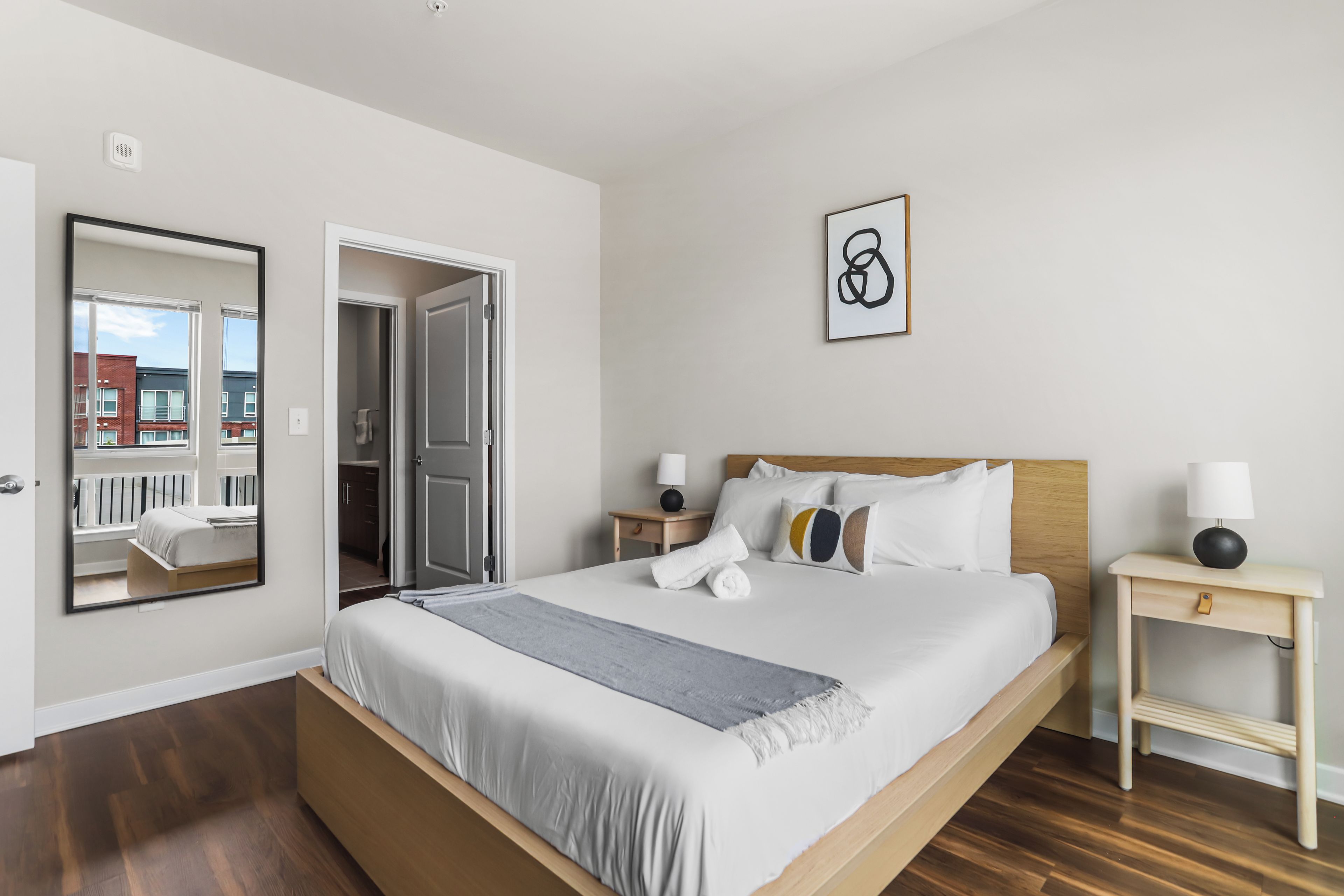 Una habitación de hotel con una cama grande, una mesita de noche con lámpara, una obra de arte en la pared y una puerta que da a un balcón.