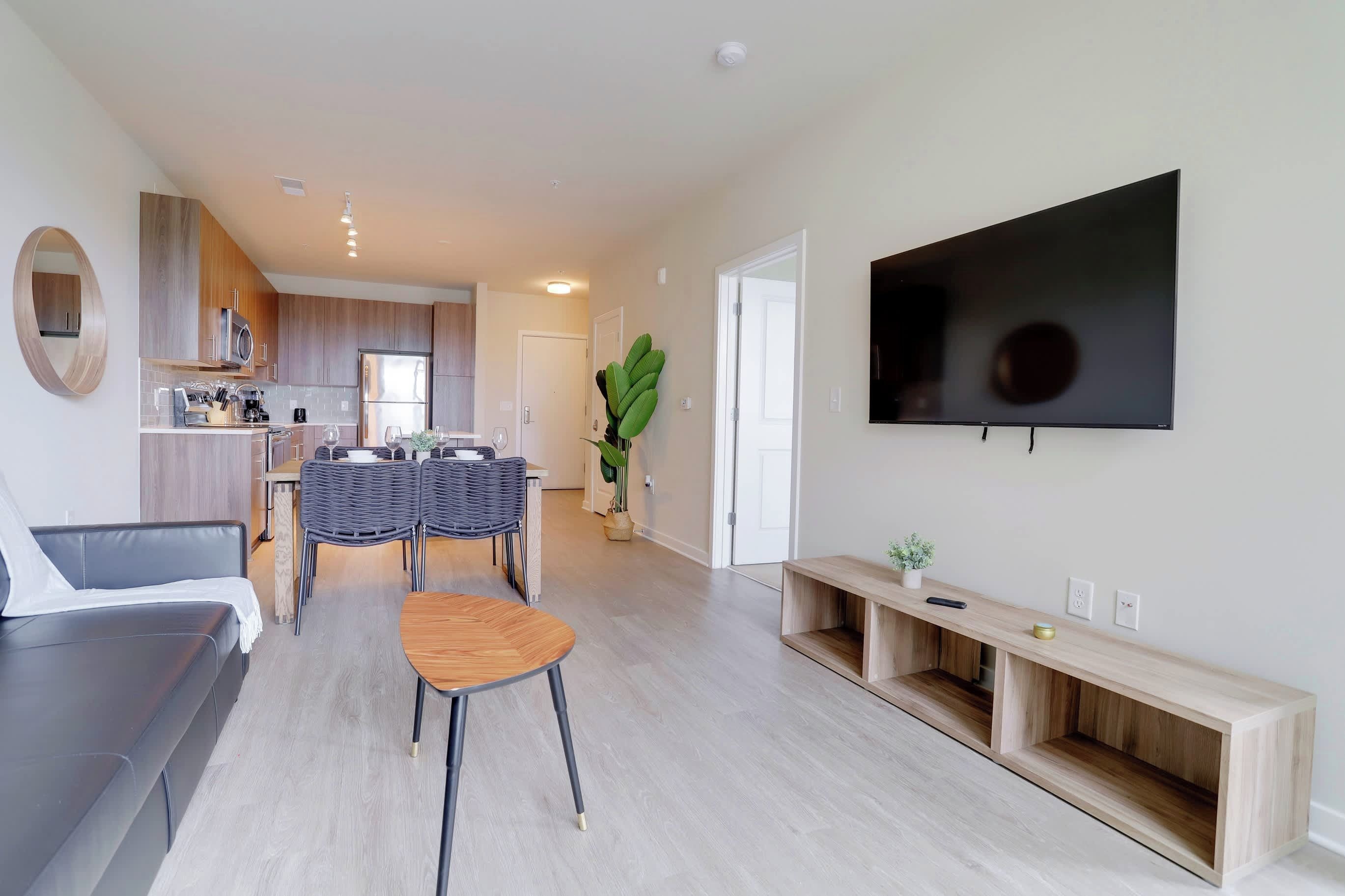 A modern living room with a sofa, a dining table with chairs, a TV mounted on the wall, and a kitchen area in the background.