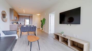 A modern living room with a sofa, a dining table with chairs, a TV mounted on the wall, and a kitchen area in the background.