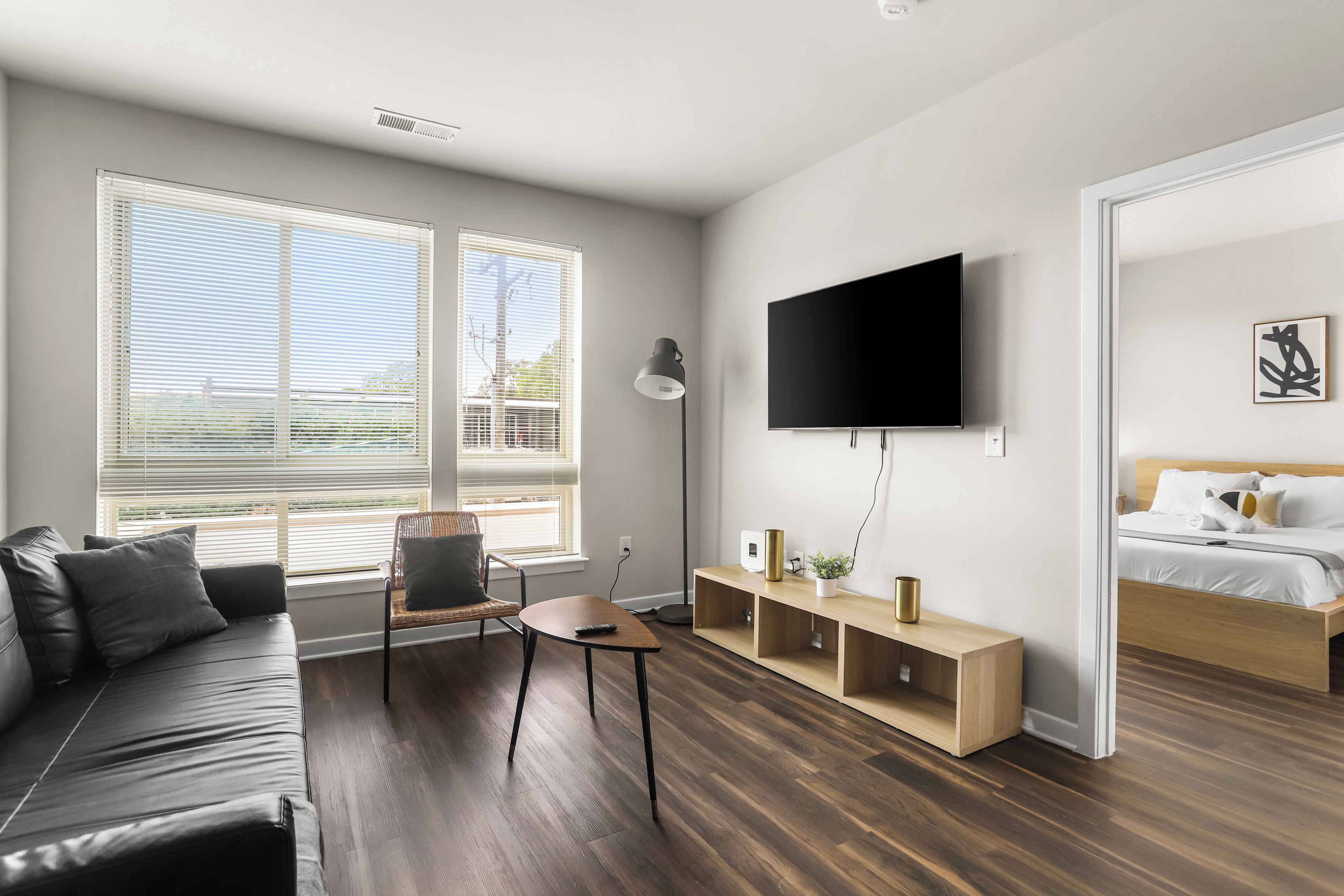 A modern living room with a sofa, a chair, a coffee table, a TV mounted on the wall, and a bedside table with a bed in the adjacent room.