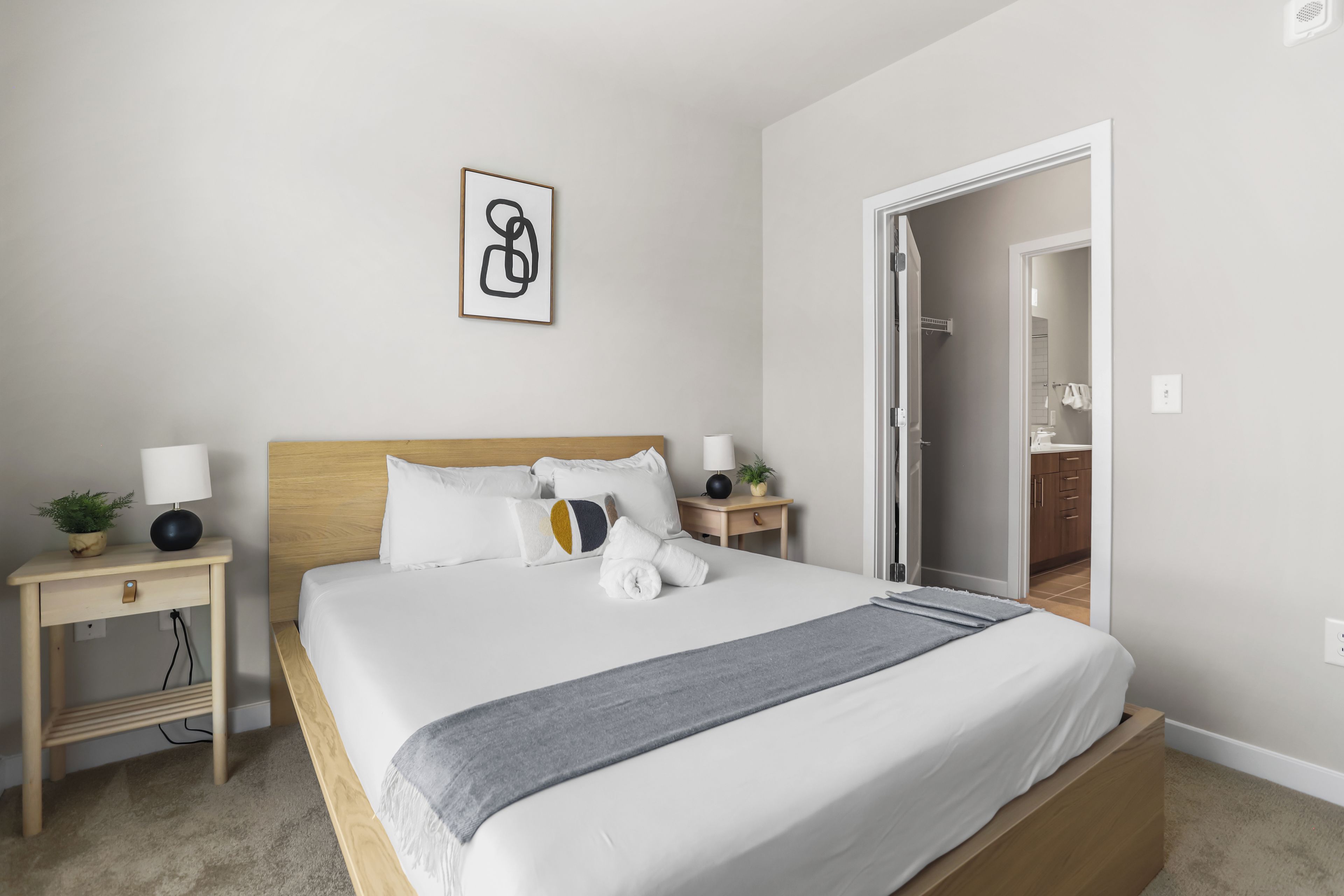 A neatly made bed with a wooden headboard, two bedside tables with lamps, a small plant, and a framed abstract artwork on the wall.